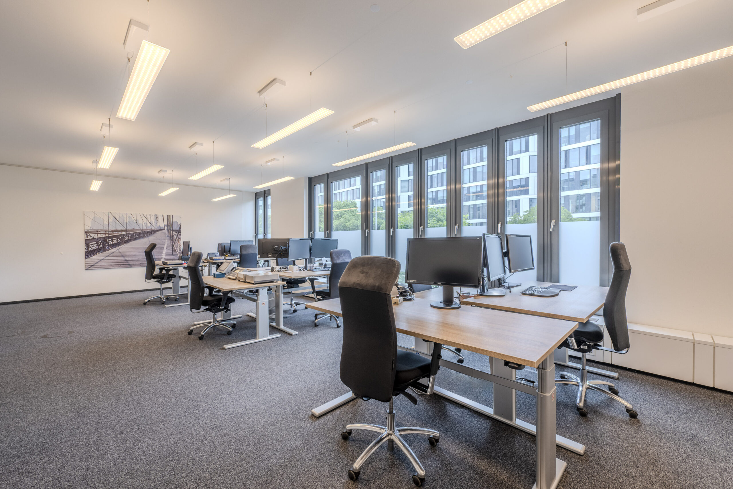 Großraumbüro mit mehreren Arbeitsplätzen und großen Fenstern.